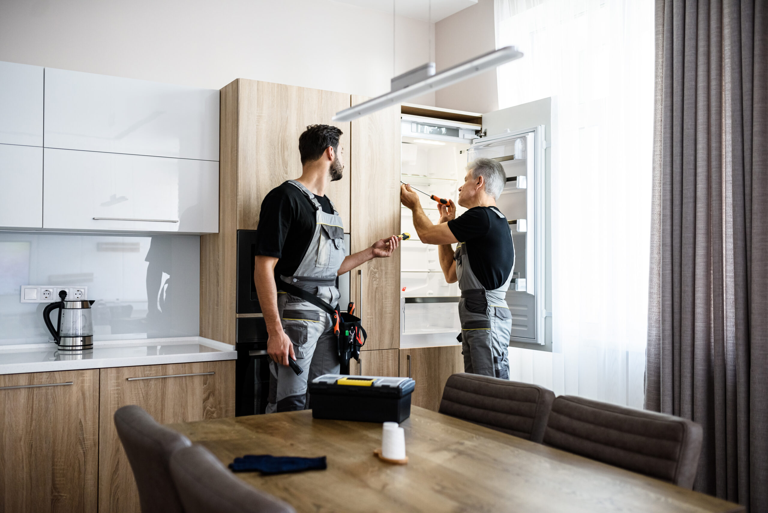 Aged repairman in uniform fixing refrigerator in the kitchen, while his colleague helping him, bringing screwdriver. Repair service concept. Selective focus. Horizontal shot